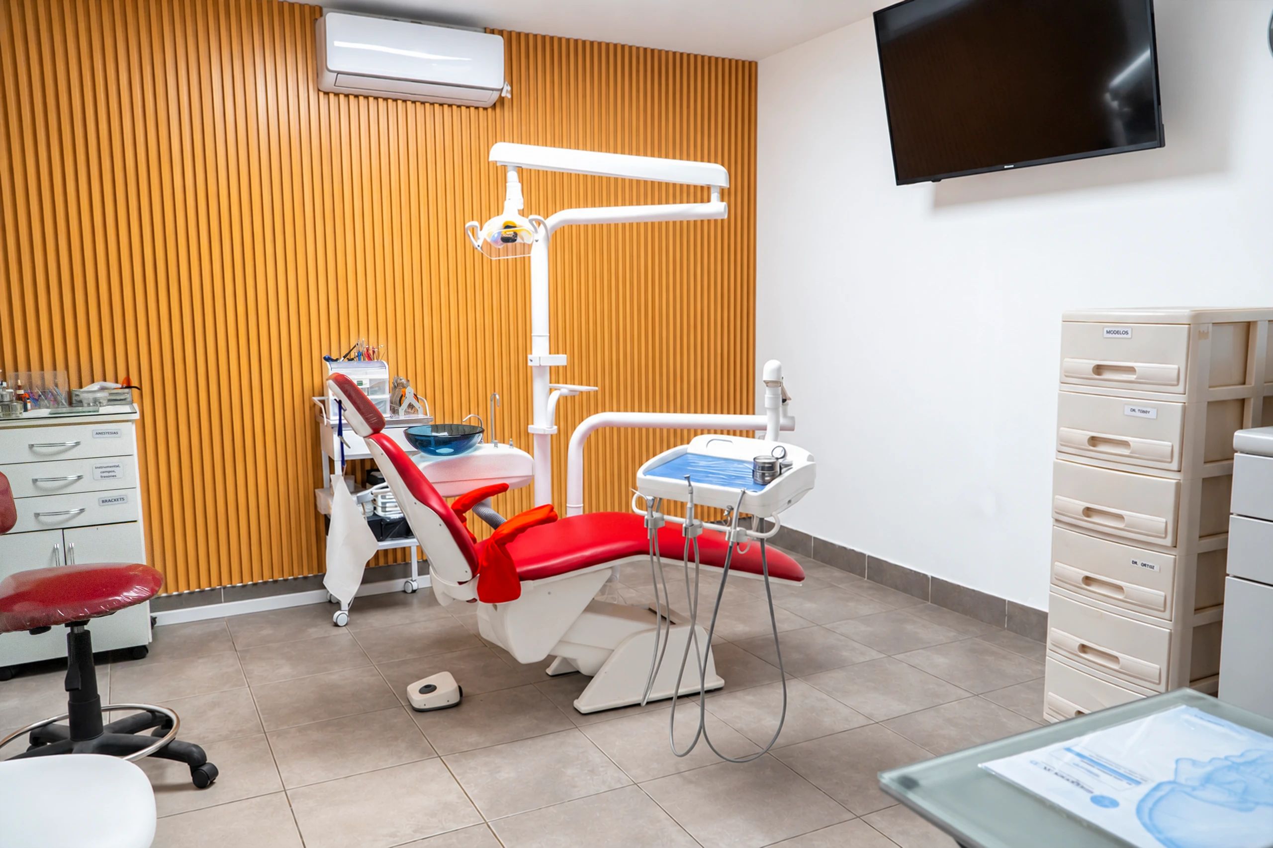 El consultorio de ortodoncia y ortopedia, con el sillón dental rojo frente a la pared revestida en madera, el televisor en la pared y los cajones de modelos a un lado