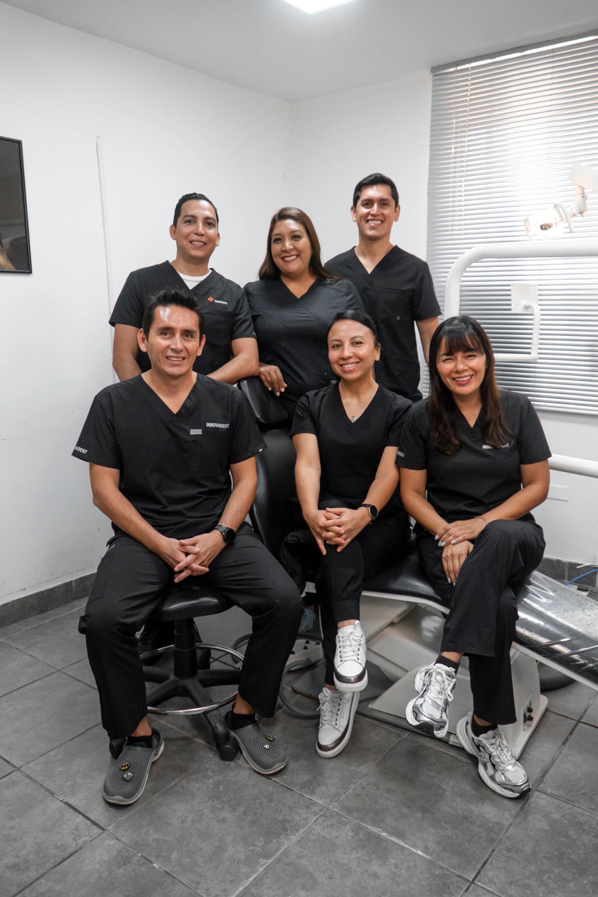 Seis especialistas de Innovadent con uniforme negro, sonriendo juntos alrededor de un sillón dental en una sala clínica: tres sentados delante y tres de pie detrás.