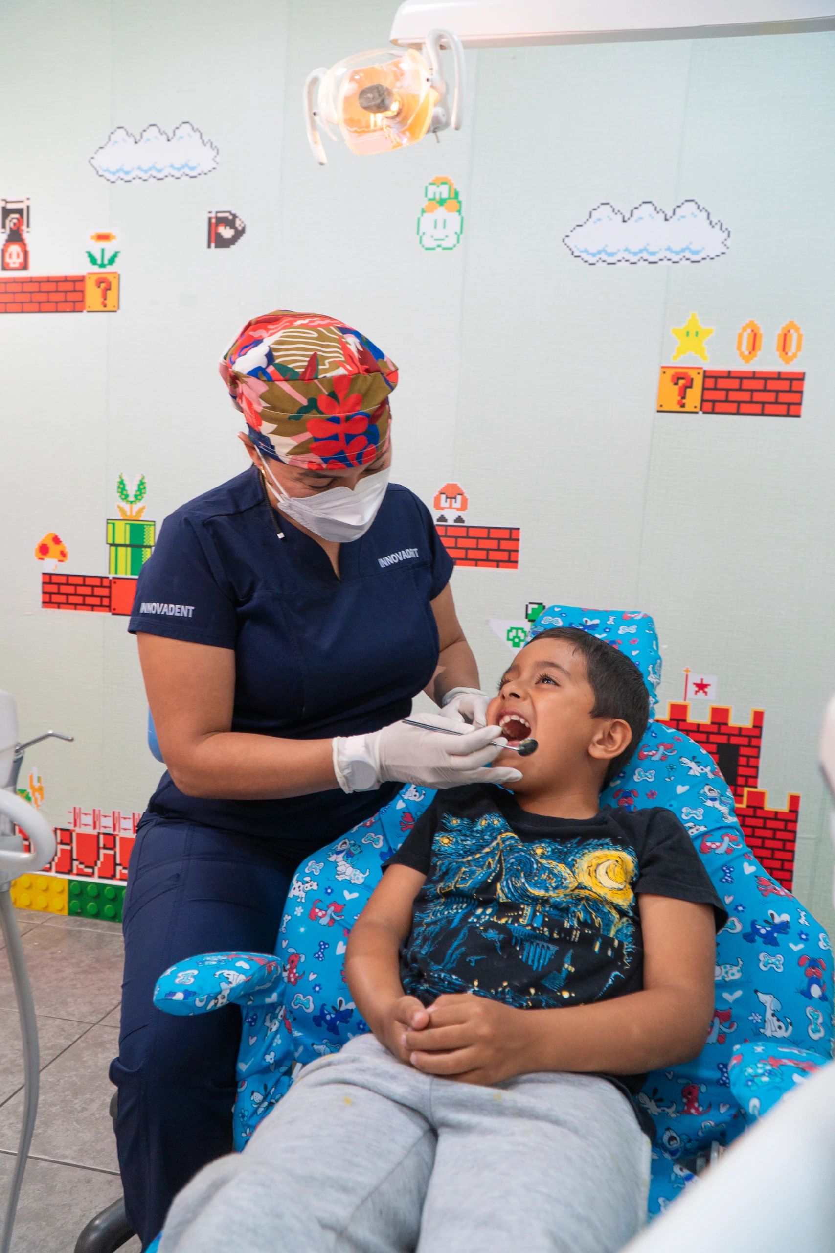 La odontopediatra revisa la boca de un niño sentado en un sillón infantil, en el consultorio decorado con figuras de videojuego.