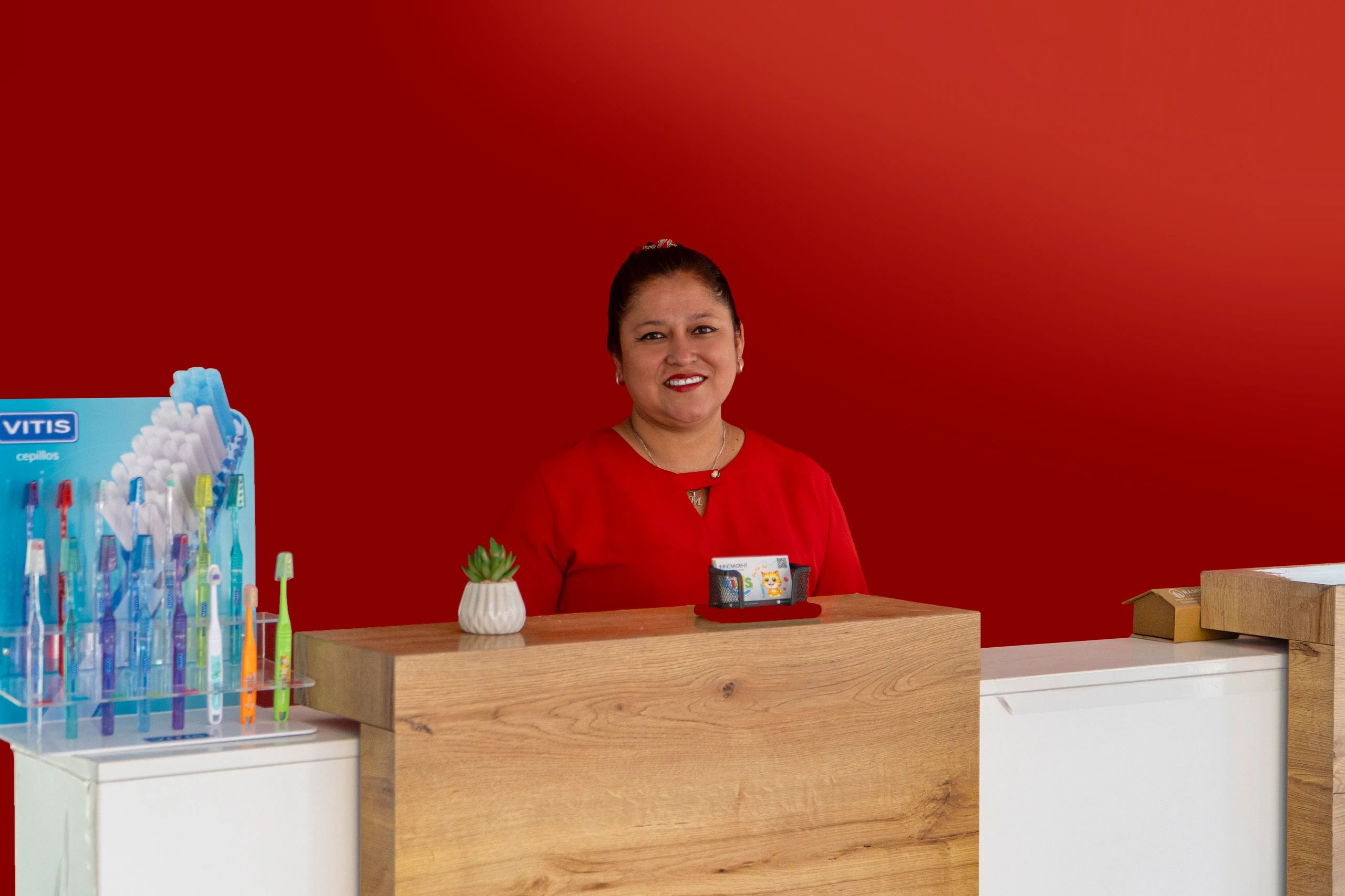 La recepcionista sonríe detrás del mostrador de madera, frente a una pared roja y junto a un exhibidor de cepillos dentales